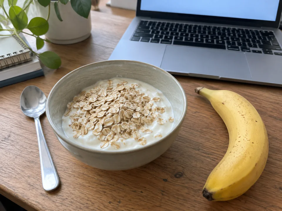 A bowl of yoghurt topped with oats and a banana beside a laptop on a working desk.